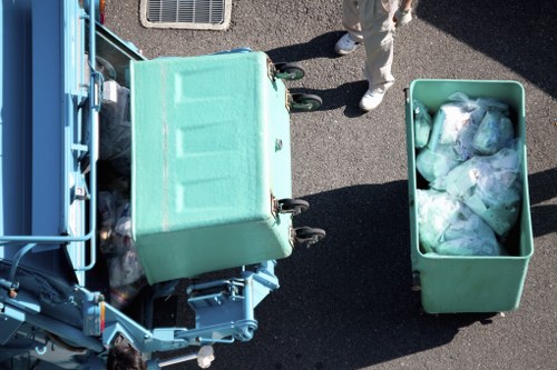 An overhead view of two large, light green waste collection bins placed on a paved surface, with one bin positioned on a waste collection vehicle and the other nearby. The bin on the vehicle appears to be filled with mixed recyclable rubbish, while the second bin contains plastic packaging and other waste materials. A person wearing light-colored trousers and shoes stands behind the bins, partially visible in the top right corner. The scene suggests a rubbish removal process possibly taking place on a driveway or street, associated with a local office clearance or waste disposal service in Eltham, London. The background shows a section of the vehicle and the surrounding pavement, emphasizing the professional handling of rubbish in a residential or commercial area.