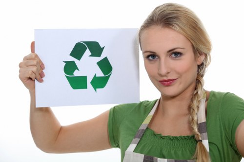 A young woman with blonde hair styled in a side braid is wearing a green top and a striped apron, standing outdoors against a plain white background. She holds a white board in her left hand, prominently displaying a green recycling symbol composed of three arrows forming a triangle. The recycling symbol signifies environmental awareness and waste management, aligning with rubbish removal services. The woman has a neutral facial expression and looks directly at the camera, conveying professionalism and responsibility. The overall composition emphasizes her association with eco-friendly waste disposal, making it relevant for a rubbish removal company's website, such as Office Clearance Eltham, serving the Eltham area, with subtle geographic relevance to London postcodes. This image visually supports themes of environmental consciousness and waste management in a professional context, without any additional props or background details visible.
