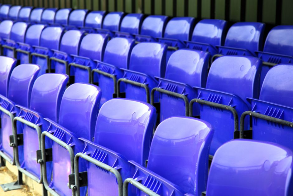 A close-up view of multiple rows of bright blue plastic stadium or auditorium seats, arranged in a uniform pattern with side-by-side placement. The seats have smooth, glossy finishes reflecting light,