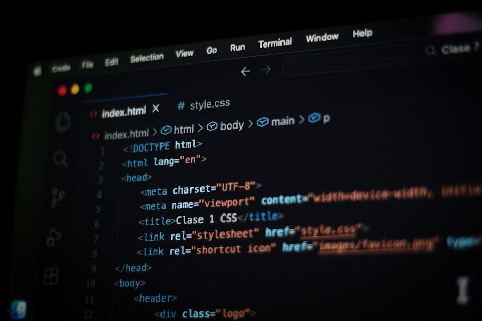 A close-up view of a person typing on a black keyboard positioned on a white desk in front of a large computer monitor displaying a code editor with lines of programming text in white and blue. The pe