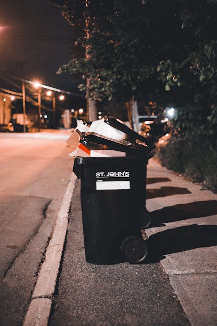 A large pile of mixed waste and rubbish is accumulated on a paved urban street corner, with an assortment of discarded cardboard boxes, plastic bags, paper, and packaging materials spilling onto the g