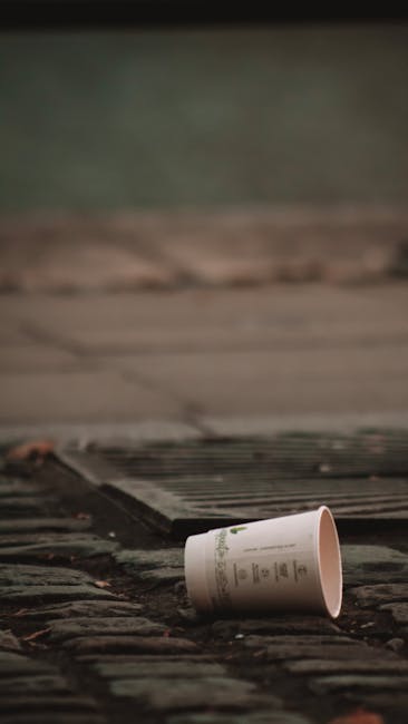 A disposable paper cup lies on its side on a rough, textured brick surface, with the opening facing towards the viewer. The cup appears to be made of light-colored paperboard with printed text and a f