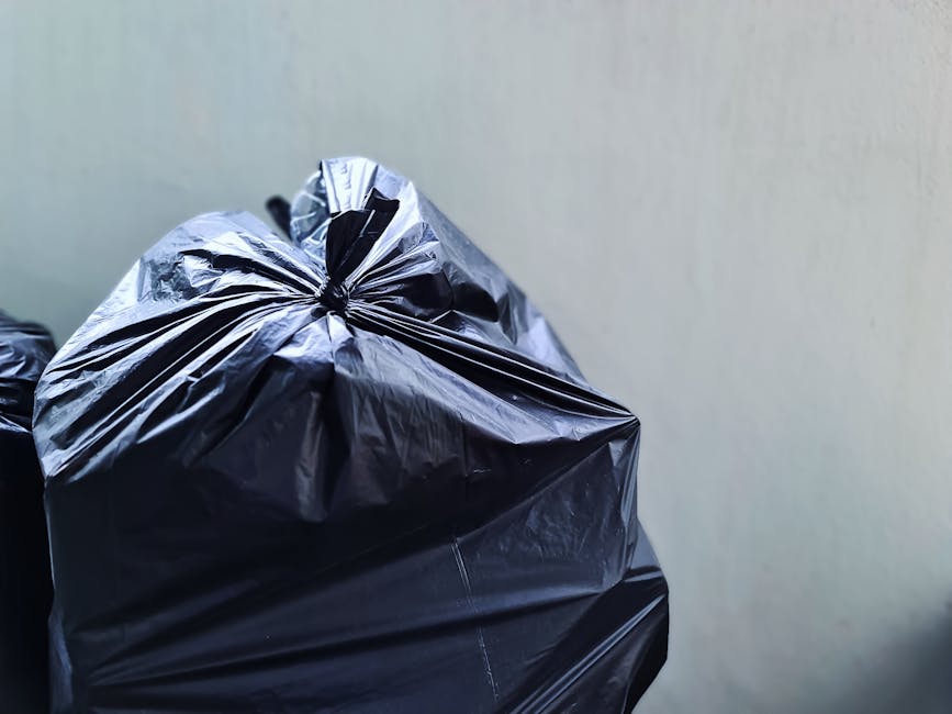 A man with short dark hair, dressed in a black t-shirt with white text on the front, is seen disposing of waste into a stainless steel litter bin while holding a large white plastic bag filled with ru