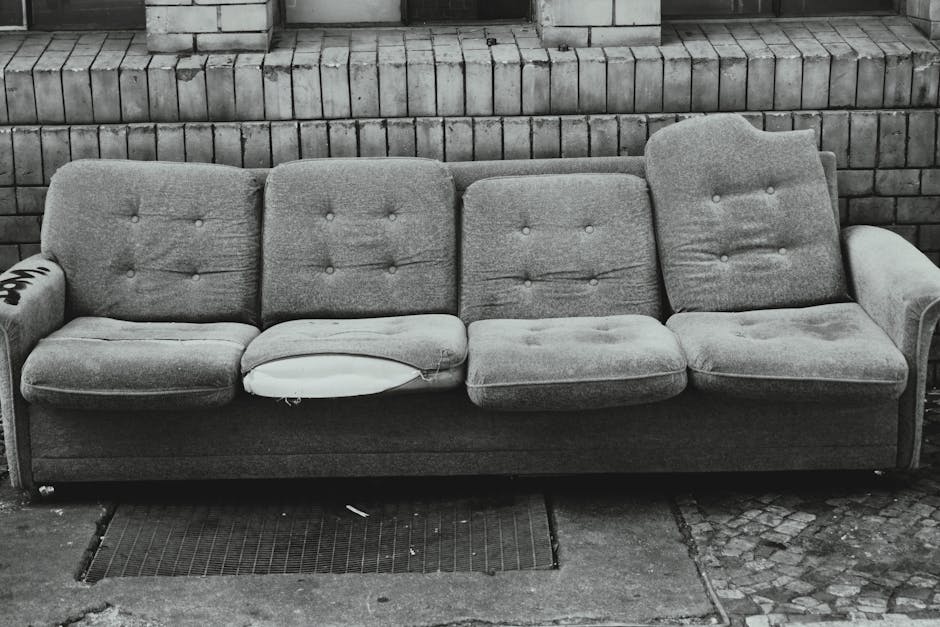 A beige upholstered armchair with padded armrests and a tufted backrest, showing visible wear and discoloration, is positioned outdoors against a weathered, graffiti-covered concrete wall. The fabric